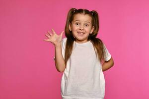 Little girl with ponytails, dressed in white t-shirt is posing against a pink studio background. Close-up shot. Sincere emotions. photo