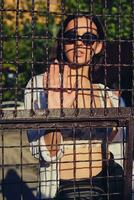 Portrait of girl in sunglasses posing in city behind a trellised fence. Dressed in top with floral print, white shirt, black trousers, waist bag. photo