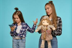 Mom and daughter dressed in checkered shirts and blue denim jeans are using smartphone while posing against a blue studio background. Close-up shot. photo