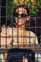 Portrait of girl in sunglasses posing in city behind a trellised fence. Dressed in top with floral print, white shirt, black trousers, waist bag. photo