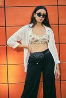 Portrait of girl in dark sunglasses posing in city against orange building. Dressed in top with floral print, white shirt, black trousers, waist bag. photo