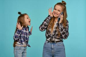 Mom and daughter dressed in checkered shirts and blue denim jeans are using smartphone while posing against a blue studio background. Close-up shot. photo