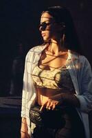 Portrait of a girl in dark sunglasses posing in city near trellised fence. Dressed in top with floral print, white shirt, black trousers, waist bag. photo