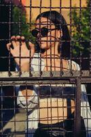 Portrait of girl in sunglasses posing in city behind a trellised fence. Dressed in top with floral print, white shirt, black trousers, waist bag. photo