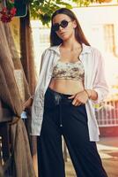 Portrait of a girl in dark sunglasses posing in city near summer terrace. Dressed in top with floral print, white shirt, black trousers, waist bag. photo