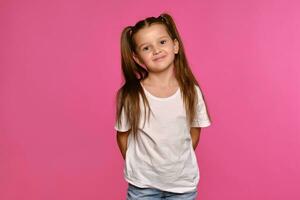 Little girl with ponytails, dressed in white t-shirt and blue jeans is posing against a pink studio background. Close-up shot. Sincere emotions. photo