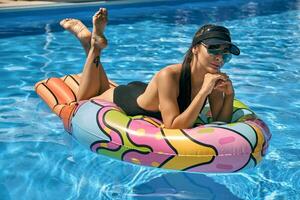 retrato de un hembra teniendo descanso y posando en un nadando piscina en un inflable colchón. vestido en un negro traje de baño, parasol gorra y Gafas de sol. foto