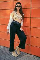 Girl in sunglasses posing in city against an orange building. Dressed in top with floral print, white shirt, black trousers, waist bag and sneakers. photo