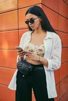 Portrait of girl in dark sunglasses posing in city against orange building. Dressed in top with floral print, white shirt, black trousers, waist bag. photo