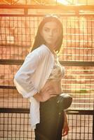 Portrait of a girl in dark sunglasses posing in city against a trellised fence. Dressed in top with floral print, white shirt, black trousers, waist bag. photo