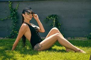 Woman posing sitting on a green grass of a recreation center or hotel with a swimming pool. Dressed in a black swimsuit, sun visor and sunglasses. photo