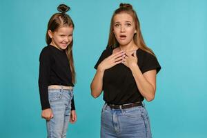 Mom and daughter with a funny hairstyles, dressed in black shirts and blue denim jeans are posing against a blue studio background. Close-up shot. photo