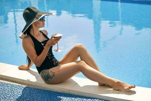 Portrait of a female having rest and posing near a swimming pool. Dressed in a black swimsuit, hat and sunglasses. photo