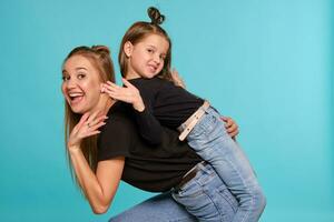 Mom and daughter with a funny hairstyles, dressed in black shirts and blue denim jeans are posing against a blue studio background. Close-up shot. photo