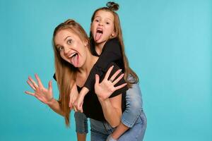 Mom and daughter with a funny hairstyles, dressed in black shirts and blue denim jeans are posing against a blue studio background. Close-up shot. photo