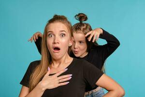 Mom and daughter with a funny hairstyles, dressed in black shirts and blue denim jeans are posing against a blue studio background. Close-up shot. photo