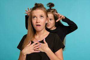 Mom and daughter with a funny hairstyles, dressed in black shirts and blue denim jeans are posing against a blue studio background. Close-up shot. photo