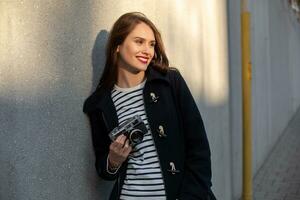 Smiling female photographer in jacket standing in front of wall ready to make new photo. Sun flare photo