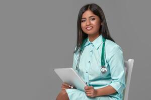 Adorable indian female doctor nurse with stethoscope in aquamarine dress sitting on chair on grey background photo