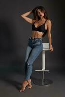 Beautiful sexy brunette woman posing in studio, sitting, looking at camera. Girl wearing fashionable jeans and sensual lingerie. photo