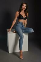 Beautiful sexy brunette woman posing in studio, sitting, looking at camera. Girl wearing fashionable jeans and sensual lingerie. photo