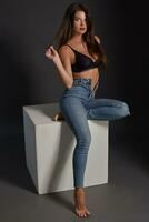 Beautiful sexy brunette woman posing in studio, sitting, looking at camera. Girl wearing fashionable jeans and sensual lingerie. photo