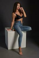 Beautiful sexy brunette woman posing in studio, sitting, looking at camera. Girl wearing fashionable jeans and sensual lingerie. photo