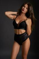 Stunning caucasian female model with dark hair and red lips in black underwear posing on dark grey background. photo