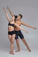 The couple of a young modern ballet dancers in black suits are posing over a gray studio background. photo