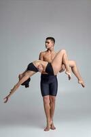 The couple of a young modern ballet dancers in black suits are posing over a gray studio background. photo