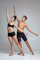 The couple of a young modern ballet dancers in black suits are posing over a gray studio background. photo