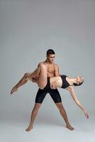 The couple of a young modern ballet dancers in black suits are posing over a gray studio background. photo