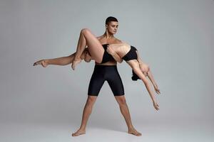 The couple of a young modern ballet dancers in black suits are posing over a gray studio background. photo