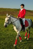 joven mujer jinete, vistiendo rojo redingote y blanco pantalones, con su caballo en noche puesta de sol ligero. foto