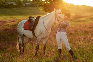 jockey joven niña caricias y abrazando blanco caballo en noche puesta de sol. Dom llamarada foto