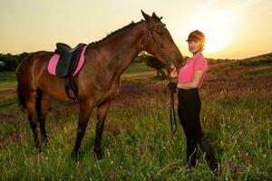 hermosa sonriente niña jockey estar siguiente a su marrón caballo vistiendo especial uniforme en un cielo y verde campo antecedentes en un puesta de sol. foto