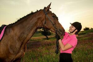 Beautiful smiling girl jockey stand next to her brown horse wearing special uniform on a sky and green field background on a sunset. photo