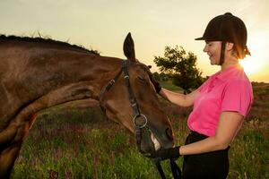 hermosa sonriente niña jockey estar siguiente a su marrón caballo vistiendo especial uniforme en un cielo y verde campo antecedentes en un puesta de sol. foto