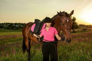 hermosa sonriente niña jockey estar siguiente a su marrón caballo vistiendo especial uniforme en un cielo y verde campo antecedentes en un puesta de sol. foto