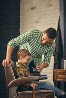 stylish little kid sitting on chair at barbershop with his young father on background photo