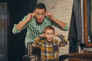 elegante pequeño niño sentado en silla a barbería con su joven padre en antecedentes foto