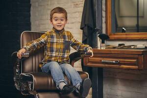 Portrait of a stylish little boy dressed in shirt and jeans in the barbershop, sitting in a chair against the barber's workplace photo