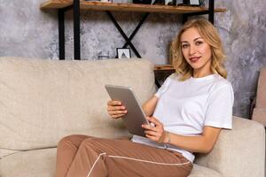 Blonde woman sitting on couch using tablet at home in the living room photo