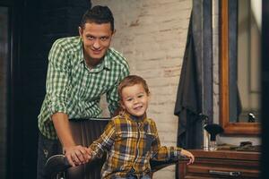 elegante pequeño niño sentado en silla a barbería con su joven padre en antecedentes foto