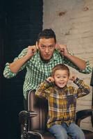 elegante pequeño niño sentado en silla a barbería con su joven padre en antecedentes foto