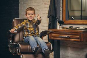 retrato de un elegante pequeño chico vestido en camisa y pantalones en el peluquería, sentado en un silla en contra el peluquería lugar de trabajo foto