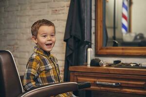 Portrait of a stylish little boy dressed in shirt and jeans in the barbershop, sitting in a chair against the barber's workplace photo