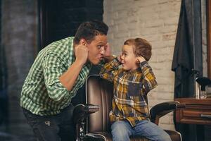 stylish little kid sitting on chair at barbershop with his young father on background photo