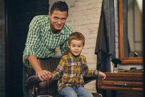 elegante pequeño niño sentado en silla a barbería con su joven padre en antecedentes foto