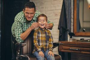 elegante pequeño niño sentado en silla a barbería con su joven padre en antecedentes foto